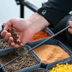 Dłoń z zanętą nad pojemnikami z różnymi składnikami do zanęty.